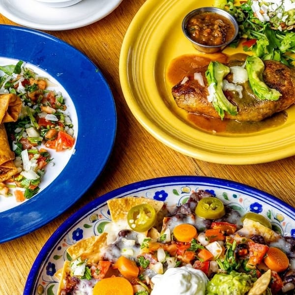 Three colorful plates of Mexican food, including taquitos, chile relleno, and nachos, on a wooden table.