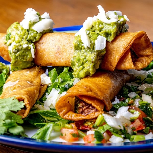 Crispy taquitos topped with guacamole, crumbled cheese, and fresh cilantro on a blue plate.