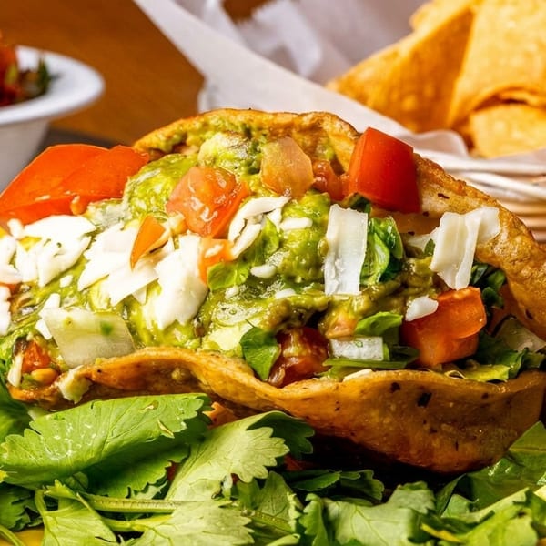 Crispy taco filled with guacamole, tomatoes, cheese, and cilantro, with salsa and chips in background.