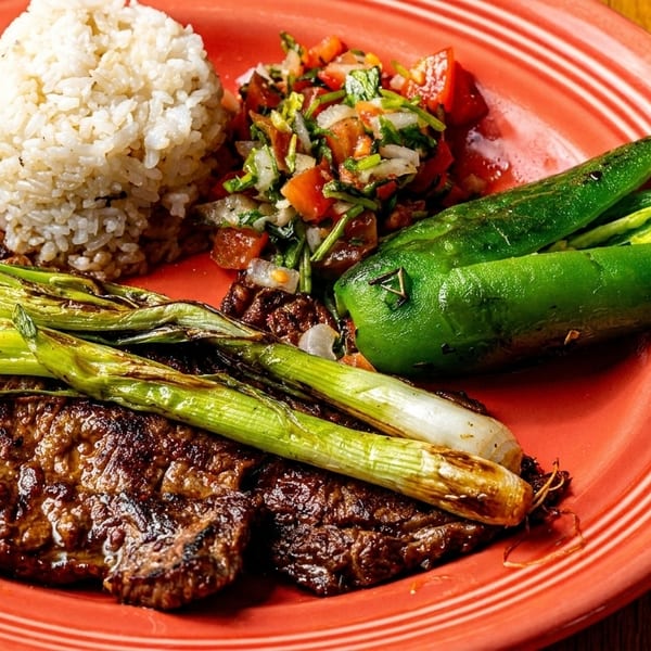 Grilled steak with green onions, rice, pico de gallo, and two green chiles on a red plate.