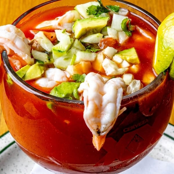 A large glass of shrimp cocktail with avocado, cucumber, and a lime wedge on a wooden table.